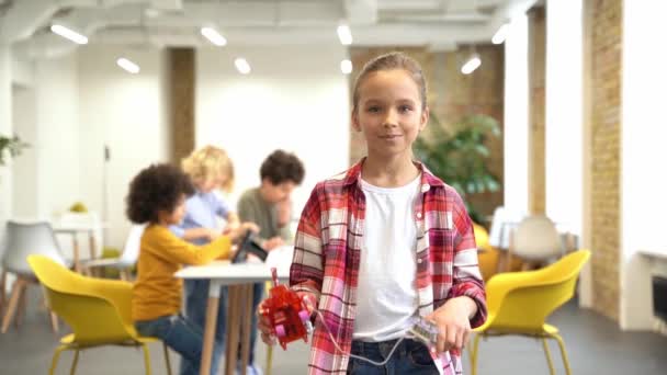 สาวน้อยที่น่ารักยิ้มที่กล้องและแสดงหุ่นยนต์ DIY ของเธอในขณะที่ยืนอยู่ในห้องเรียนระหว่างบทเรียน STEM วีดีโอสต็อกที่ปลอดค่าลิขสิทธิ์