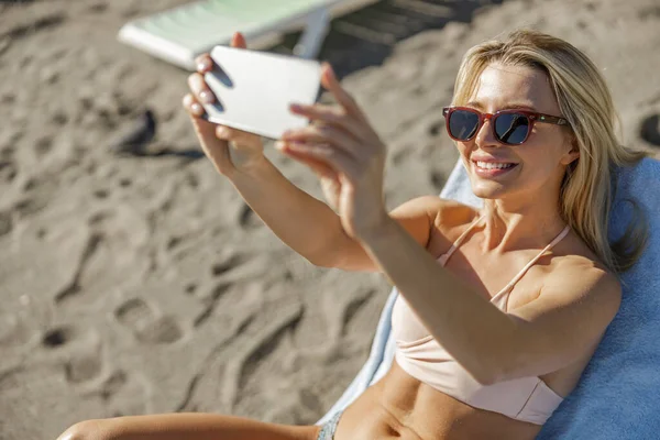 Tatilde gülümseyen genç bayan sahilde dinlenirken akıllı telefon kullanarak fotoğraf çekiyor.