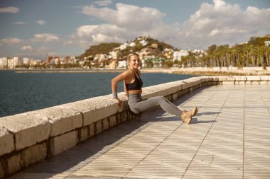 Gülümseyen genç sporcu kadın deniz kenarında ısınma egzersizi yapıyor.