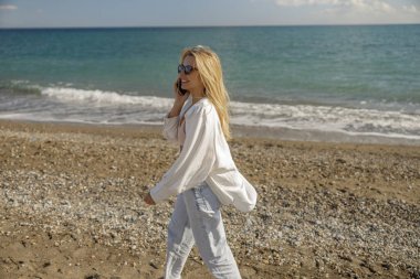 Güneş gözlüklü kadın telefonla konuşuyor. Kumsalda tek başına yürürken akıllı telefon kullanıyor.