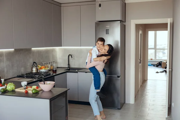 Cheerful young pretty mother holding small teen son on hands in kitchen and laughing. — Fotografia de Stock