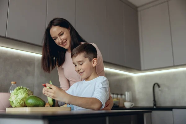 Cheerful young Caucasian mother with small son peeling vegetables and cooking. — Foto de Stock
