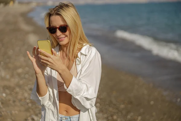 Mujer joven en gafas de sol usando la aplicación móvil mientras pasa tiempo en una playa — Foto de Stock