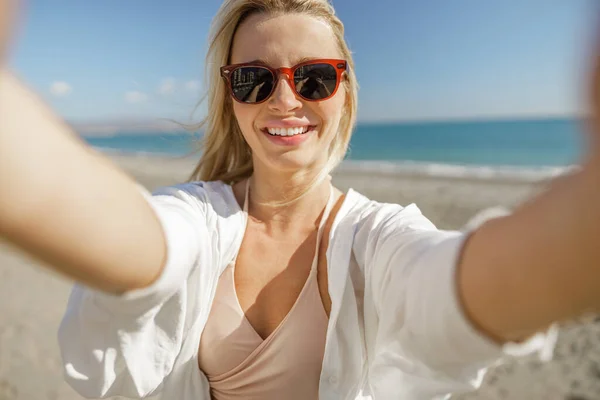 Cute blonde woman wearing sunglasses making selfie on the beach — Stock fotografie