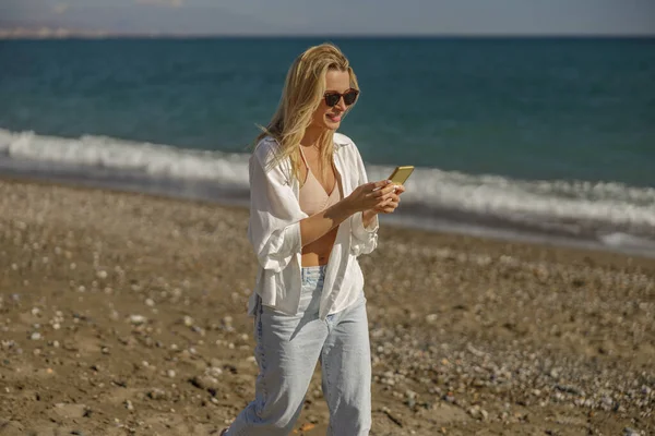Sahilde yürürken akıllı telefon kullanarak mesajlaşan odaklanmış bir kadın.
