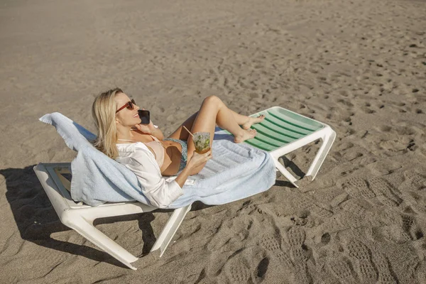 Güverte sandalyesinde uzanmış telefonla konuşuyor ve sahilde kokteyl içiyor.