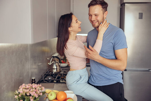 Joyful young Caucasian man and woman laughing and hugging in kitchen in the morning.