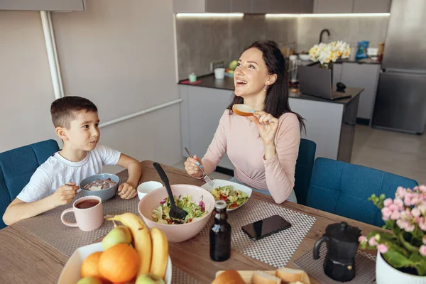 Cheerful mother eating salat in the morning and small son having breakfast with cereals. — Foto de Stock