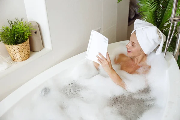 Top view on young Caucasian pretty female relaxing in bathtub with foam and reading book. — Stok Foto
