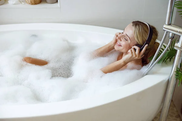Vrolijke jonge vrouw in koptelefoon glimlachend, luisteren naar muziek in bad met bubbels. — Stockfoto