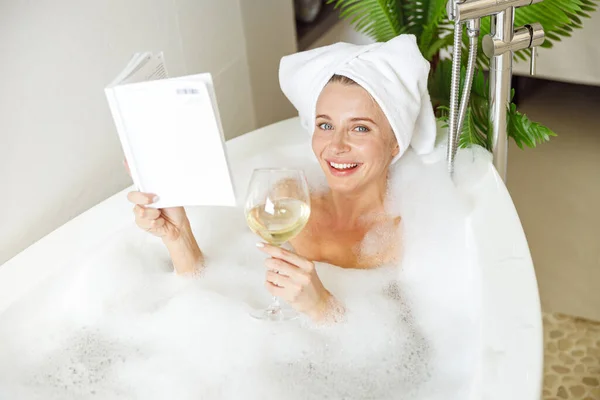 Portrait of pretty smiled young female lying in bath with foam, reading and drinking wine. — Stockfoto