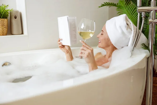 Pretty young relaxed female with towel on head taking bath, drinking wine and reading. — Fotografia de Stock