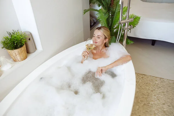 Blonde young attractive female lying in bubbles in bathtub and drinking white wine. — Photo