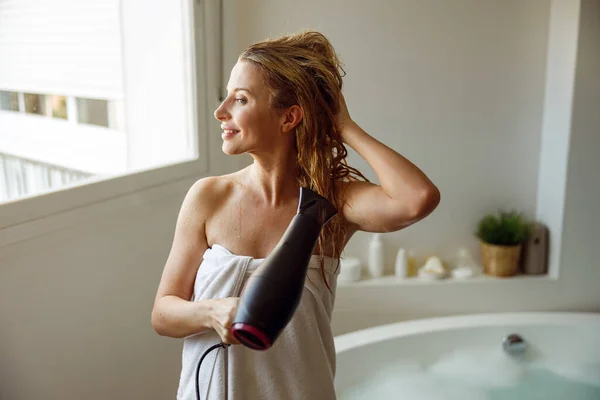 Joyful young Caucasian pretty female drying hair with dryer after taking foam bath. — ストック写真