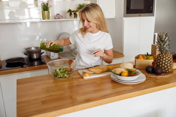 Güzel sarışın kadın mutfaktaki yeşil salataya zeytin yağı ekliyor. Sağlıklı yaşam tarzı. — Stok fotoğraf