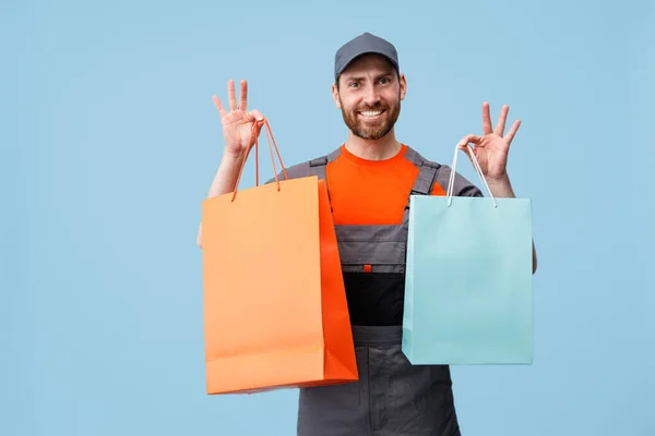 Happy delivery man in uniform holding shopping bags on blue studio background — Stock Fotó