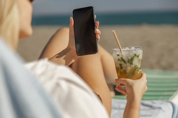 Primer plano de las manos femeninas con smartphone y cóctel en un día de playa soleado — Foto de Stock