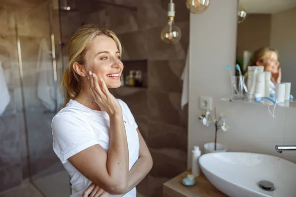 Wanita muda pirang yang mempesona tersenyum di cermin di kamar mandi. Konsep perawatan diri. — Stok Foto
