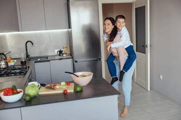 Retrato de lindo niño pequeño sentado en la espalda de mamá bastante feliz y sonriendo en la cocina. — Foto de Stock