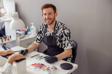 Plastik eldiven ve önlük giyen neşeli bir manikür uzmanı kuafördeki kadın müşteriye manikür yapıyor.