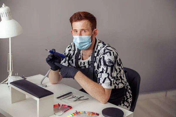 Confident nail master wearing protective mask and gloves holding electric nail file drill in hand while sitting at working table