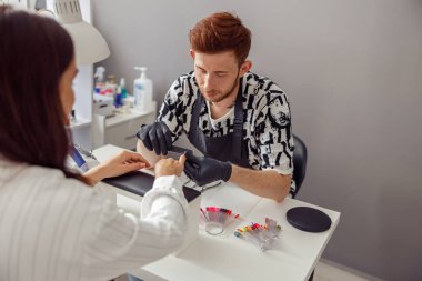 Profesyonel manikür ustası, siyah lateks eldiven ve önlük giyiyor. Modern kuaförde tırnak törpüsüyle müşteri tırnakları üzerinde çalışıyor.