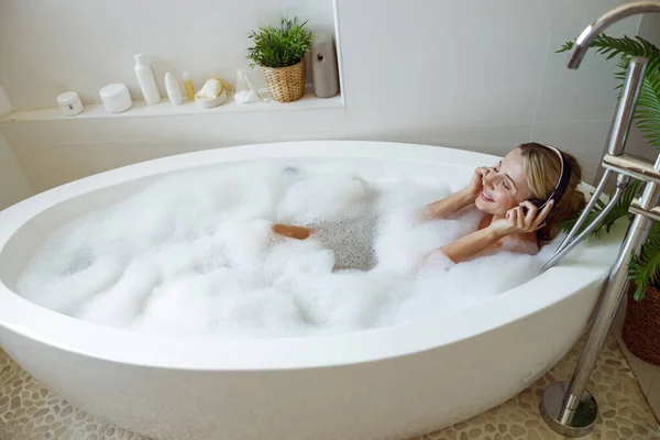 泡風呂に寝そべって音楽を聴いているヘッドフォンの白人の若いきれいな女性 トップ表示 — ストック写真