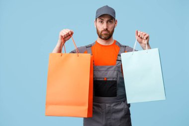 Mavi arka planda alışveriş torbaları tutan üniformalı ciddi bir teslimatçı. Stüdyo çekimi