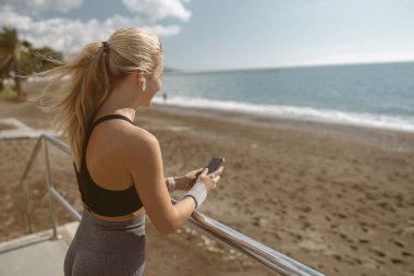 Kablosuz kulaklık takan kadın koşucunun arka planı. Kıyı şeridindeki trabzanlar ve akıllı telefonları tararken spor aktivitelerinden dinleniyor.