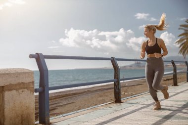 Plajda koşarken gülümseyen spor sutyen ve tayt giyen sağlıklı genç kadının tam boy portresi.