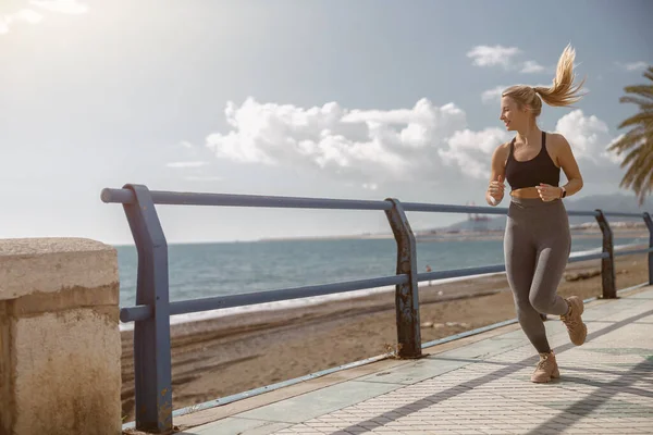 Plajda koşarken gülümseyen spor sutyen ve tayt giyen sağlıklı genç kadının tam boy portresi.