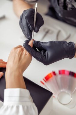 Lastik eldivenli çivi ustasının ellerinde kesilmiş. Kuaför salonunun bulanık arka planında metal manikür aletiyle tırnak eti itiyor.