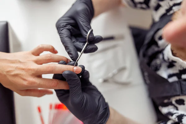 Cropped on hands of nail technician wearing protective gloves using nail scissors for working with clients manicure in beauty shop