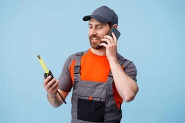 Üniformalı bir adam cep telefonuyla konuşuyor ve elinde mavi stüdyonun arka planında mezura var.. 