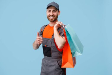 Üniformalı gülümseyen erkek kurye alışveriş çantası tutuyor ve mavi stüdyo arka planında baş parmağını gösteriyor..