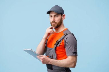 Üniformalı bir erkek işçi mavi stüdyo arka planında elinde tabletle sakalına dokunuyor.