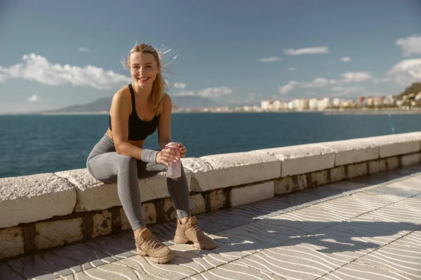 Spor elbiseli güzel bayan su şişesi tutuyor ve deniz kenarında koşmaktan yorulduktan sonra dinleniyor.