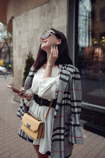 Beautiful smiled woman in stylish outlook standing at city street with smartphone in hand. Fashion.