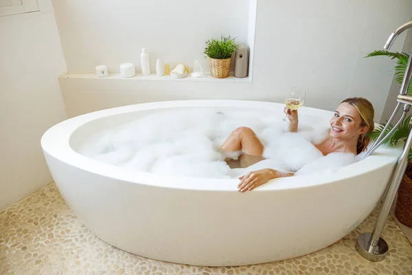 泡の入った浴槽に寝そべって白ワインのグラスを持っている金髪の美しい若い女性 肖像画 — ストック写真