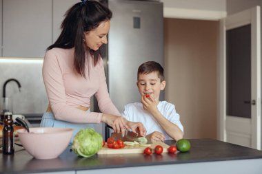 Kafkasyalı mutlu anne, sebze doğruyor, küçük tatlı oğlu vişneli domates tadıyor ve yiyor.