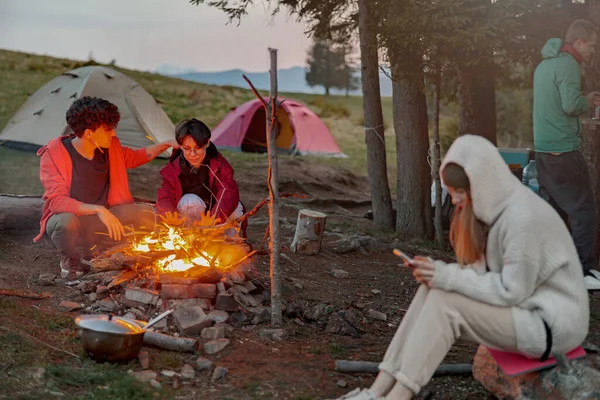 Caucasian friends hikers warming up at fire in camp site in mountains. Evening. Vacation in nature.