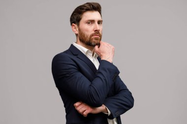 Ellerinin çapraz olduğu klasik siyah takım elbiseli bir iş adamı gri stüdyo arka planındaki kameraya bakıyor.