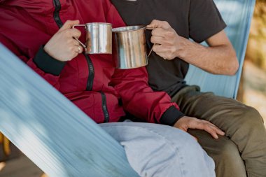 Close up of steel cups of coffee or tea in hands of man and woman in hammock. Rest in forest. Cheers