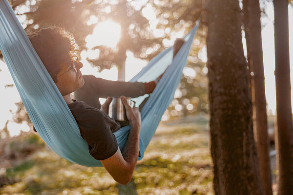 Handsome young man in glasses lying in hammock and relaxing with coffee. Forest travel. Rest in wood