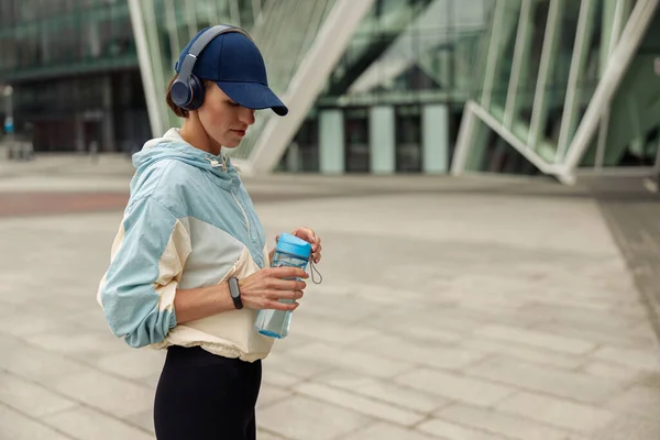Woman in headphones stands with bottle of water after running on background of modern building.