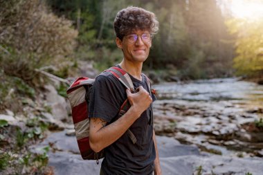 Portrait of young cheerful male tourist with backpack standing at river in mountains and smiling. 