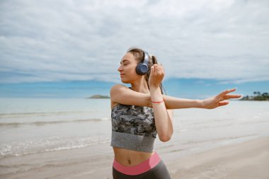 Sportive woman in headphone making stretching exercises on beach. Healthy and active life concept