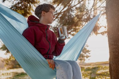 Young woman in glasses sitting in hammock with thermos in forest and enjoying morning. Rural tourism