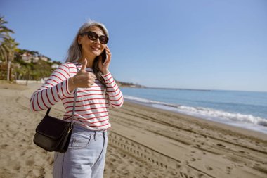 Rahat, olgun bir kadın, güneşli bir günde kumsalda dikilirken cep telefonuyla konuşuyor ve baş parmağını bırakıyor.