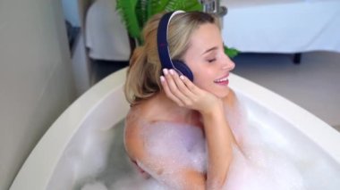 Genç mutlu sarışın kadın köpükle banyo yapıyor ve kulaklıkla müzik dinliyor. Üst görünüm.